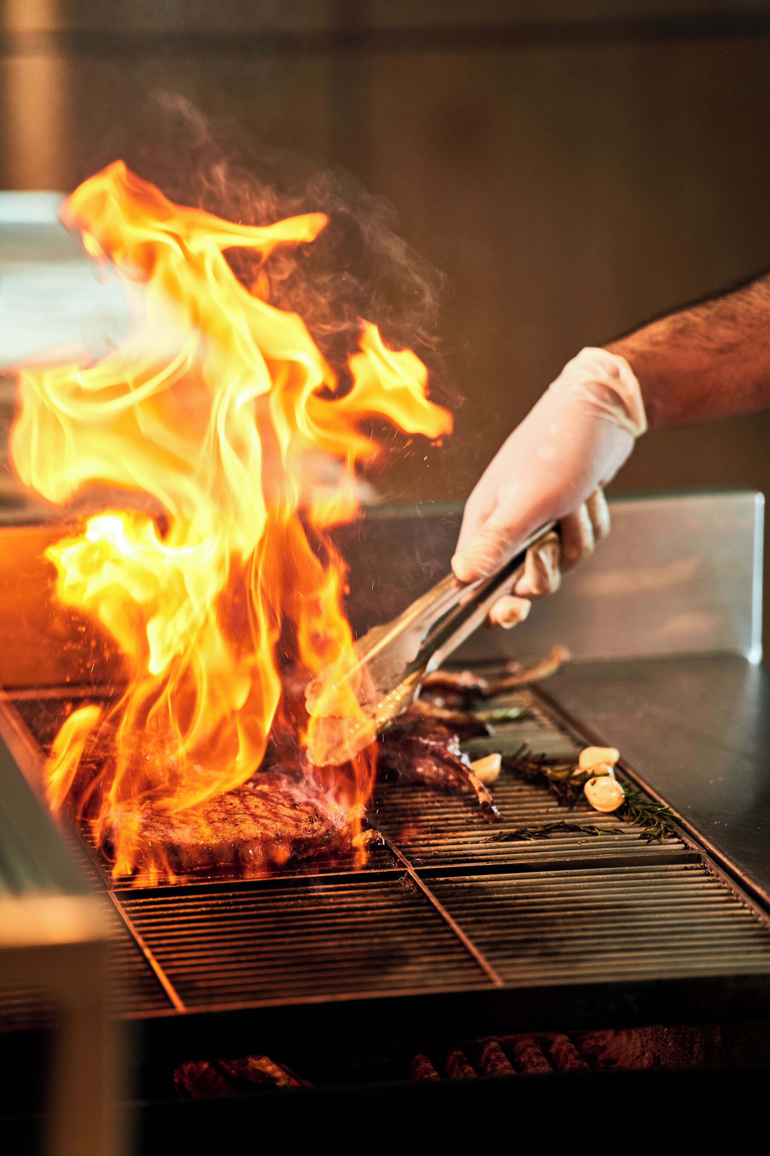 A grill with flames and a gloved hand holding tongs.