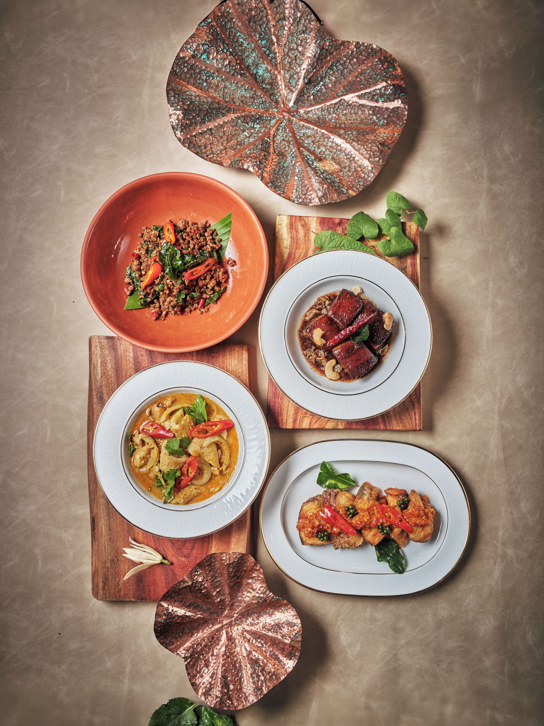 A top-down view of a dining table with four plates of food, each on a wooden board, and a copper leaf decoration.