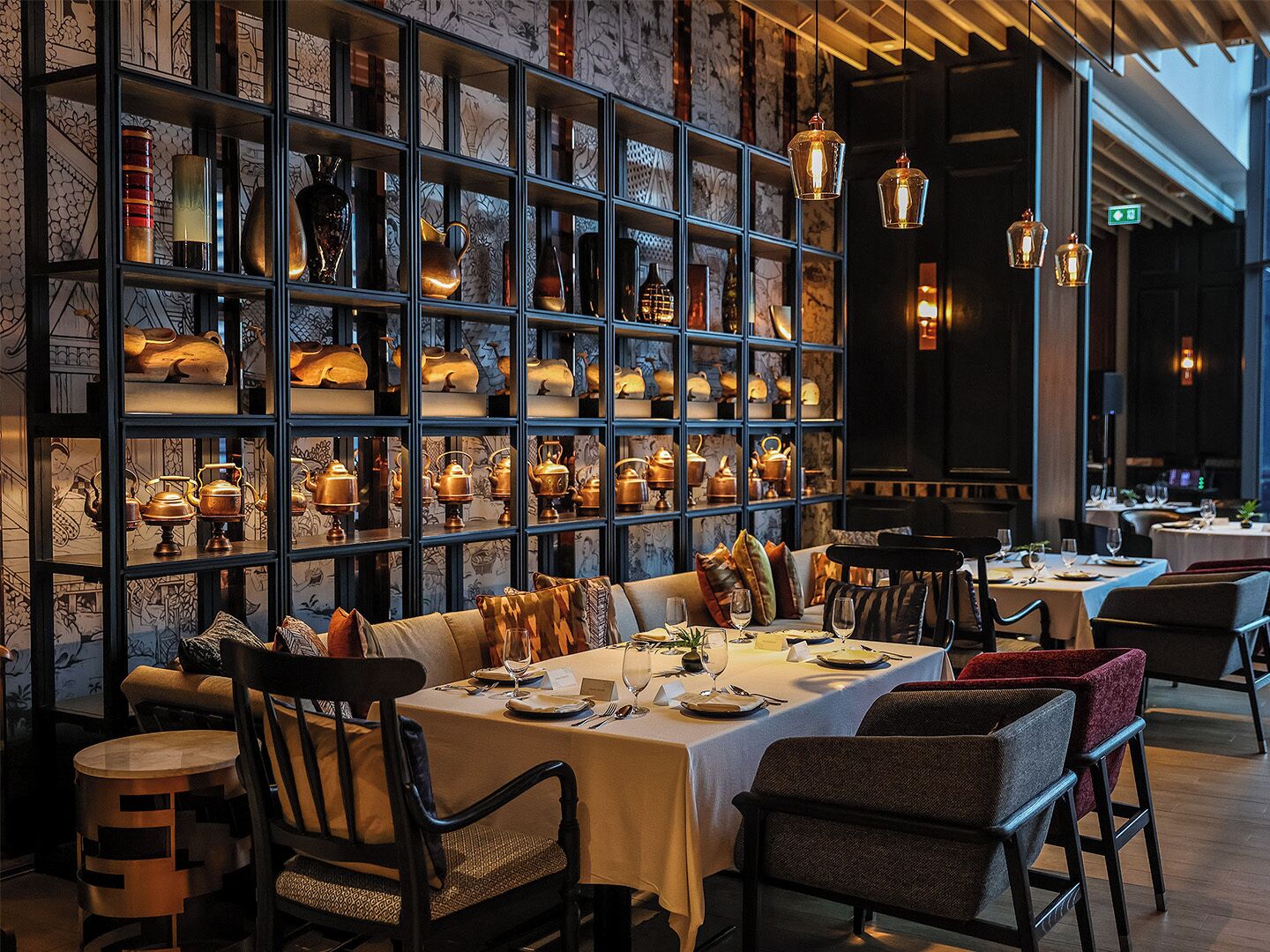 A restaurant with a long dining table, multiple chairs, and a wall of shelves displaying various dishes and pottery.