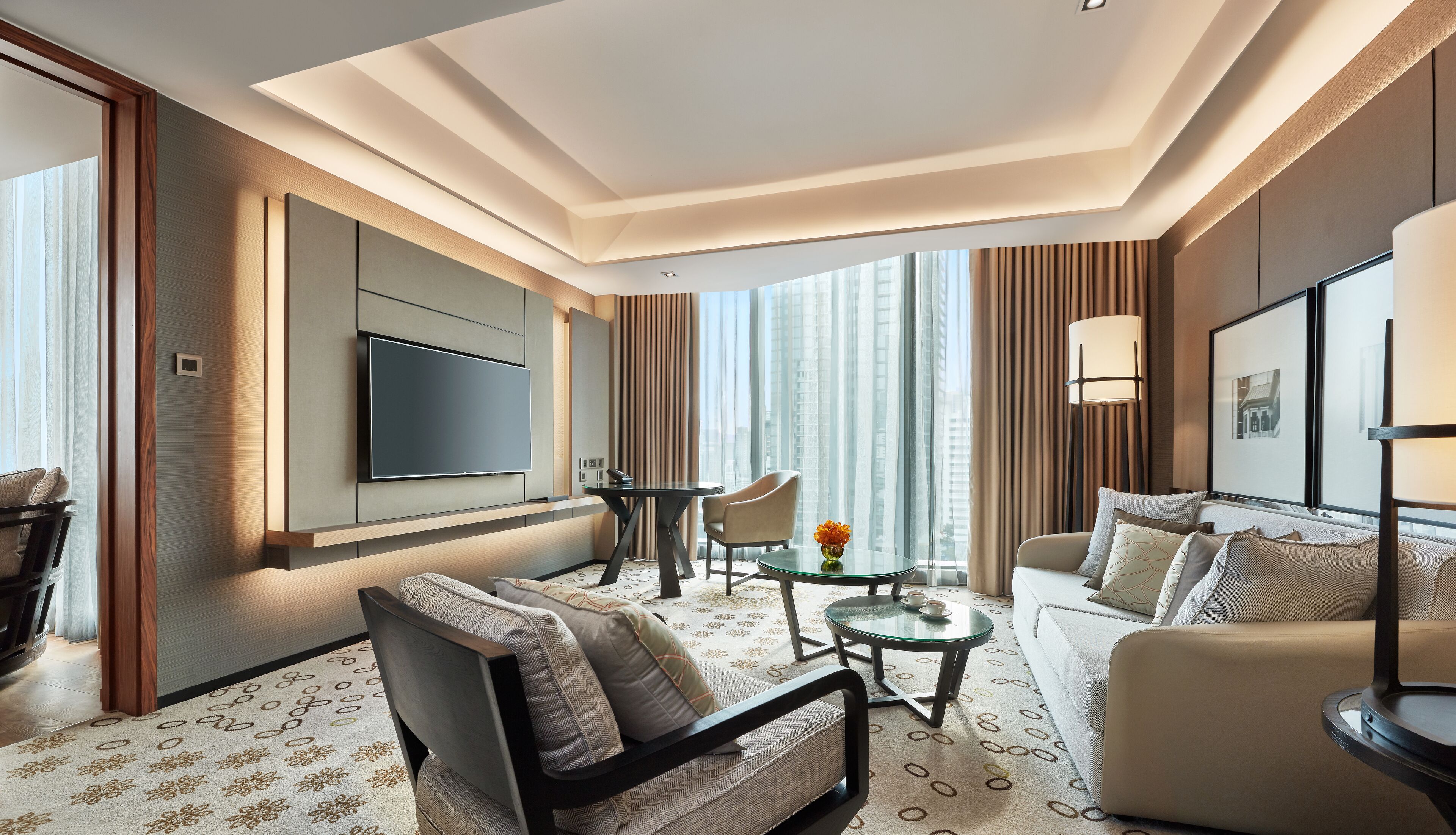 A modern hotel room with a flat-screen TV, a sofa, a chair, a small table with a vase, and a large window with curtains.