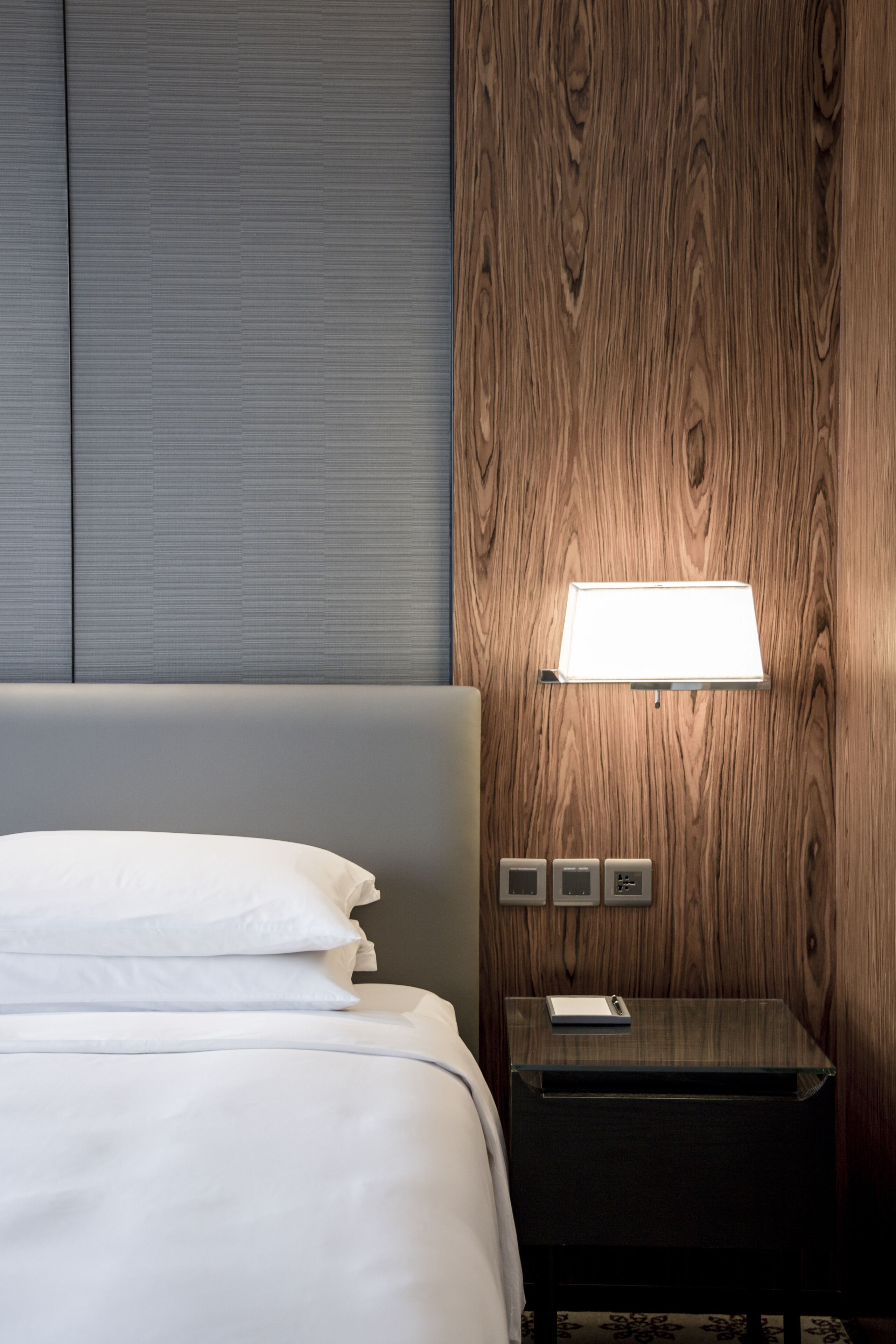 A neatly made bed with white linens, a bedside table with a lamp, and a wall with a wooden panel design.