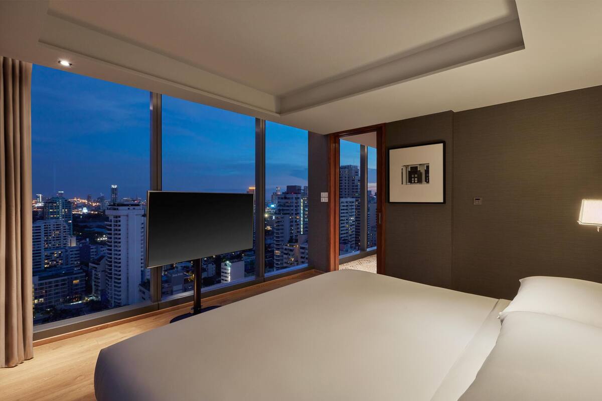 Une chambre d’hôtel avec un grand lit, un téléviseur à écran plat, un tableau encadré au mur et une vue sur le paysage urbain à travers de grandes fenêtres.