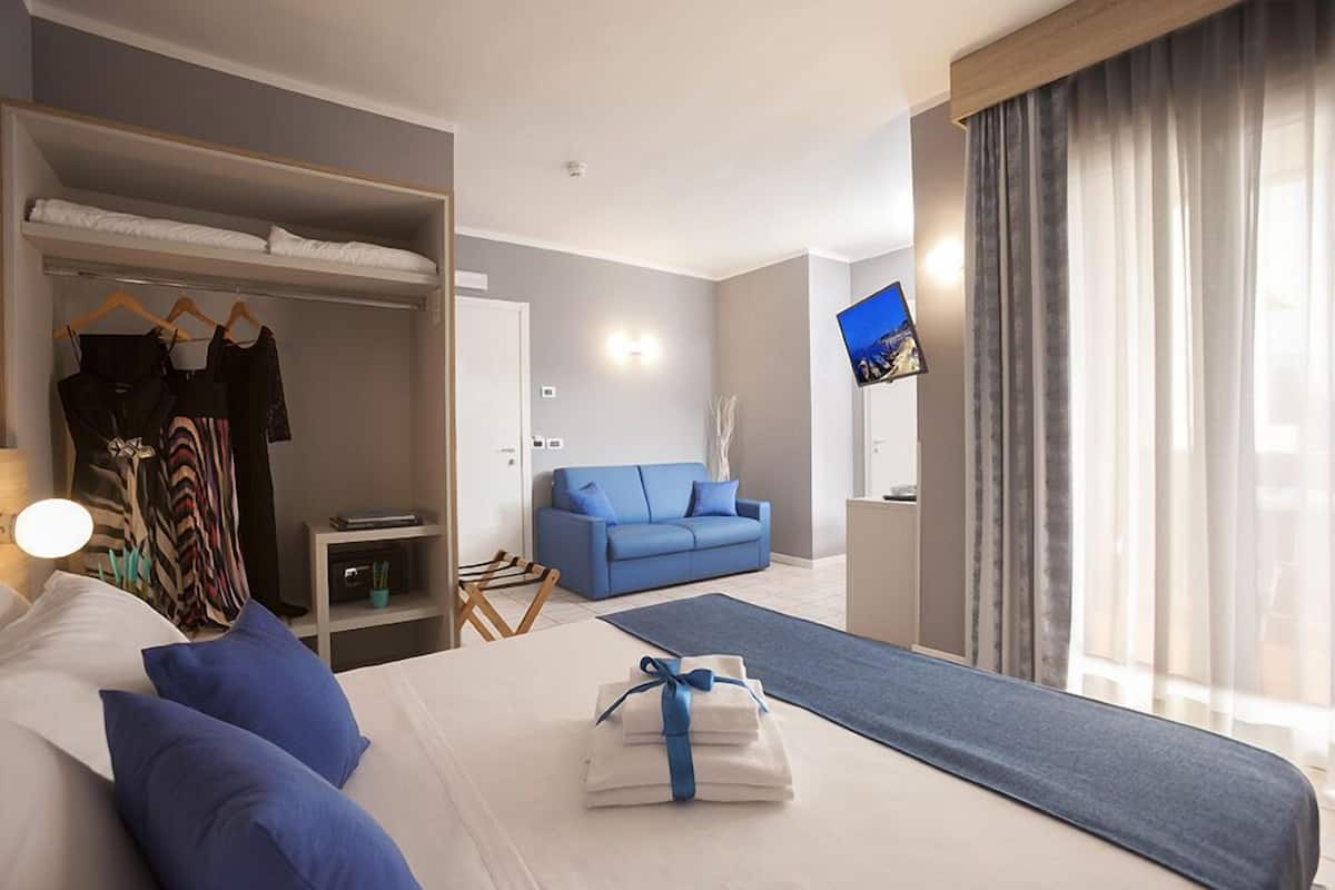 A hotel room with a bed, a blue sofa, a wardrobe, a TV, and a window with curtains.