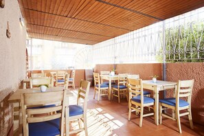 An outdoor patio area with wooden tables and chairs, a tiled floor, and a view of greenery through a metal fence.