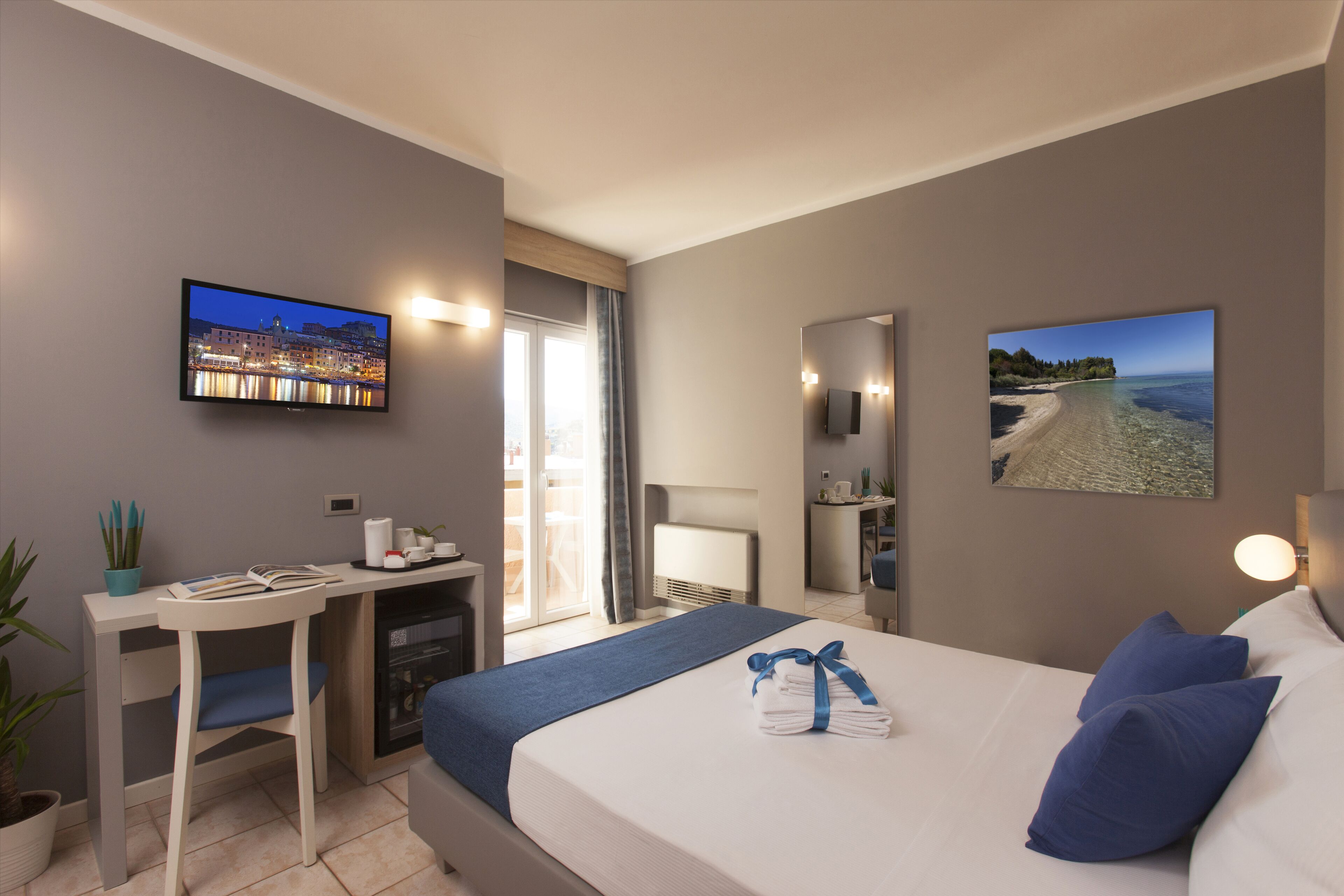 Une chambre d’hôtel équipée d’un lit, d’un bureau, d’une télévision et d’un balcon offrant une vue sur le paysage urbain.