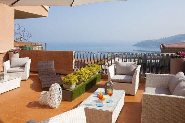A terrace with wicker furniture, a small table with drinks, and a view of the sea.