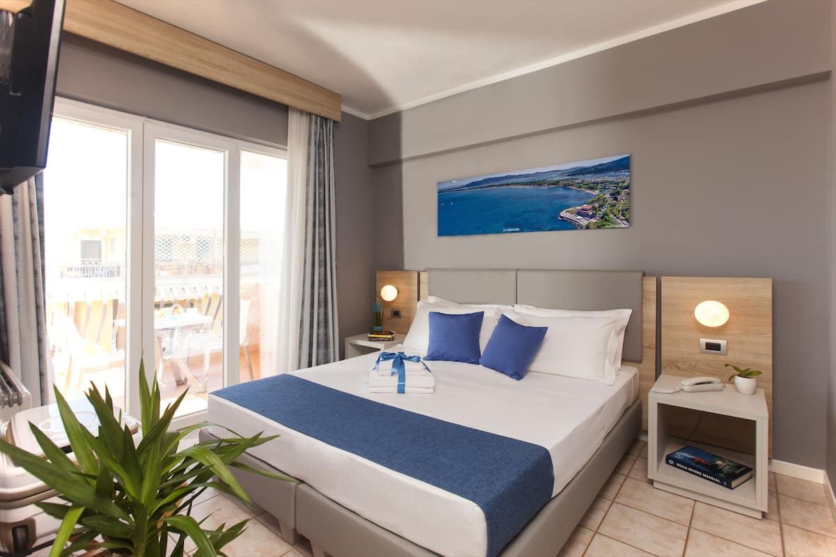 A hotel room with a large bed, a view of the sea, a painting on the wall, and a small plant on the nightstand.