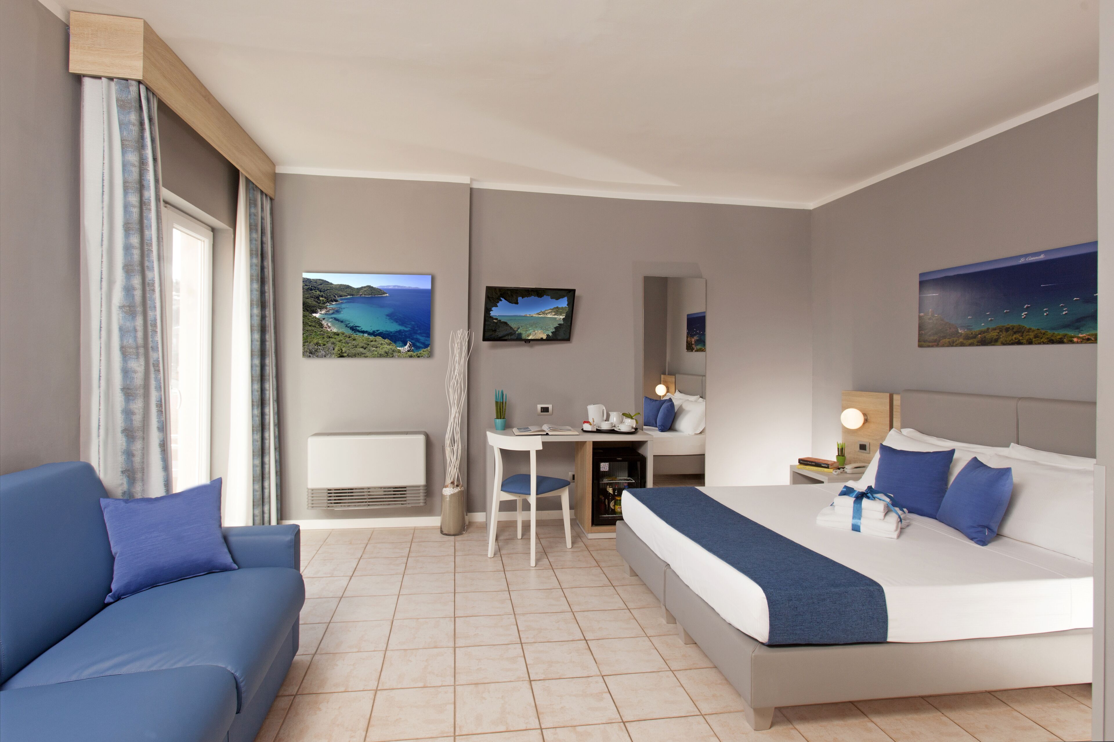 A hotel room with a bed, a desk, a chair, a sofa, and two wall-mounted TVs.