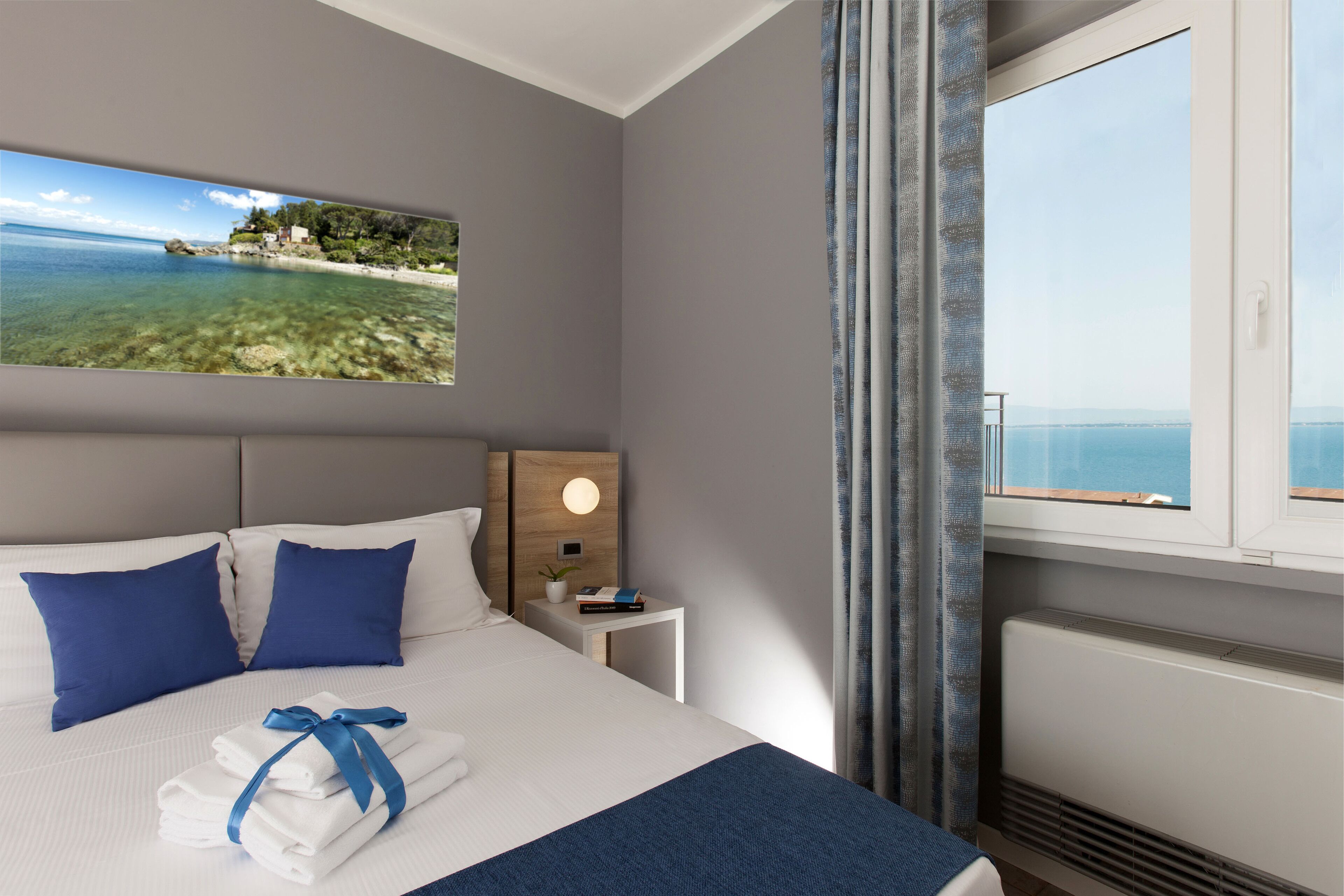 Una cama bien hecha con ropa de cama azul y blanca, una mesita de noche con una lámpara y vista al mar a través de una ventana.