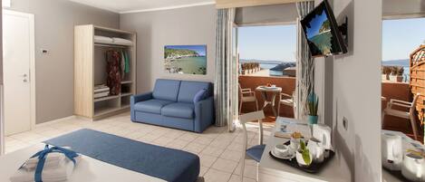 A hotel room with a bed, a blue sofa, a dining area with a table and chairs, a TV mounted on the wall, and a balcony with a view of the sea.