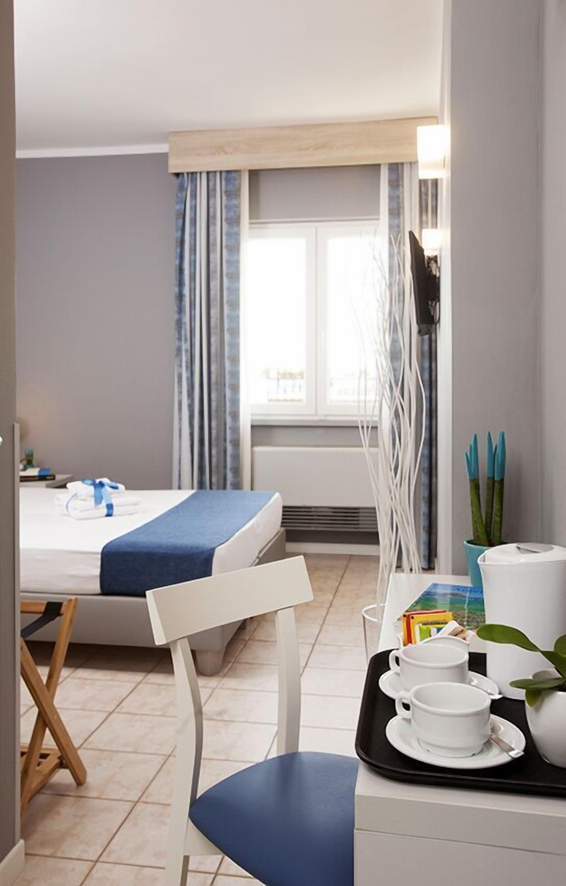 Een hotelkamer met een bed, een eettafel met een dienblad vol witte kopjes en schotels, een stoel, en een raam met gordijnen.