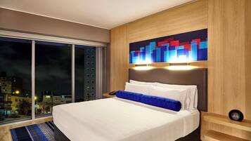 A modern hotel room with a large bed, a city view, and a wooden headboard.