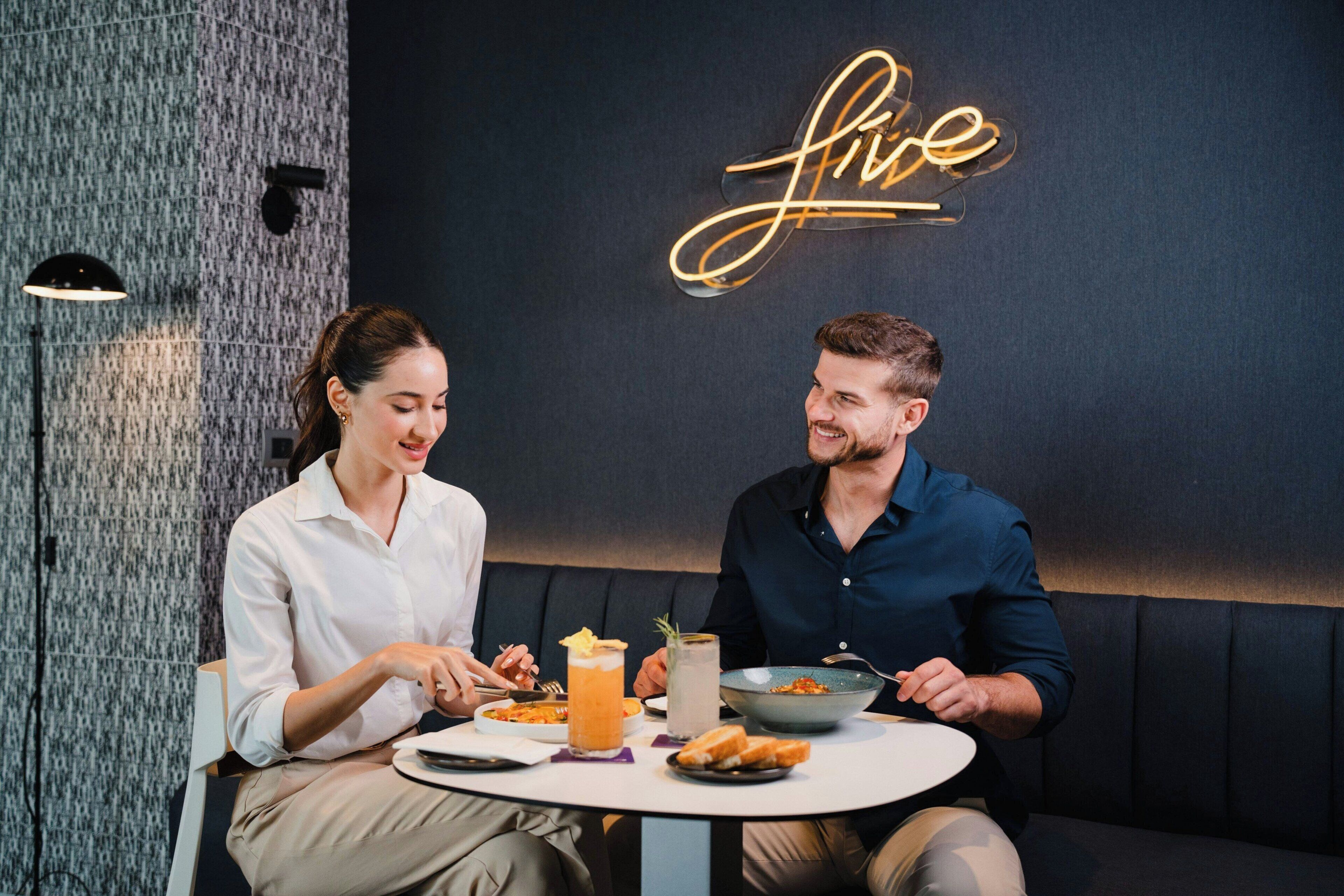 Dos personas cenando en una mesa, con un letrero de neón al fondo.