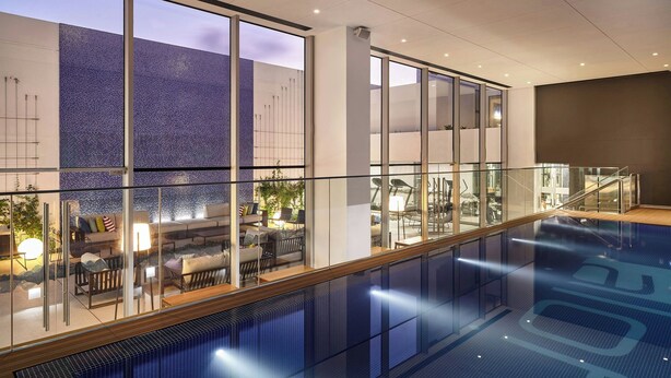 Een modern binnenzwembad met glazen balustrades, een zithoek met een bank en stoelen, en uitzicht op een muur met blauwe tegels.