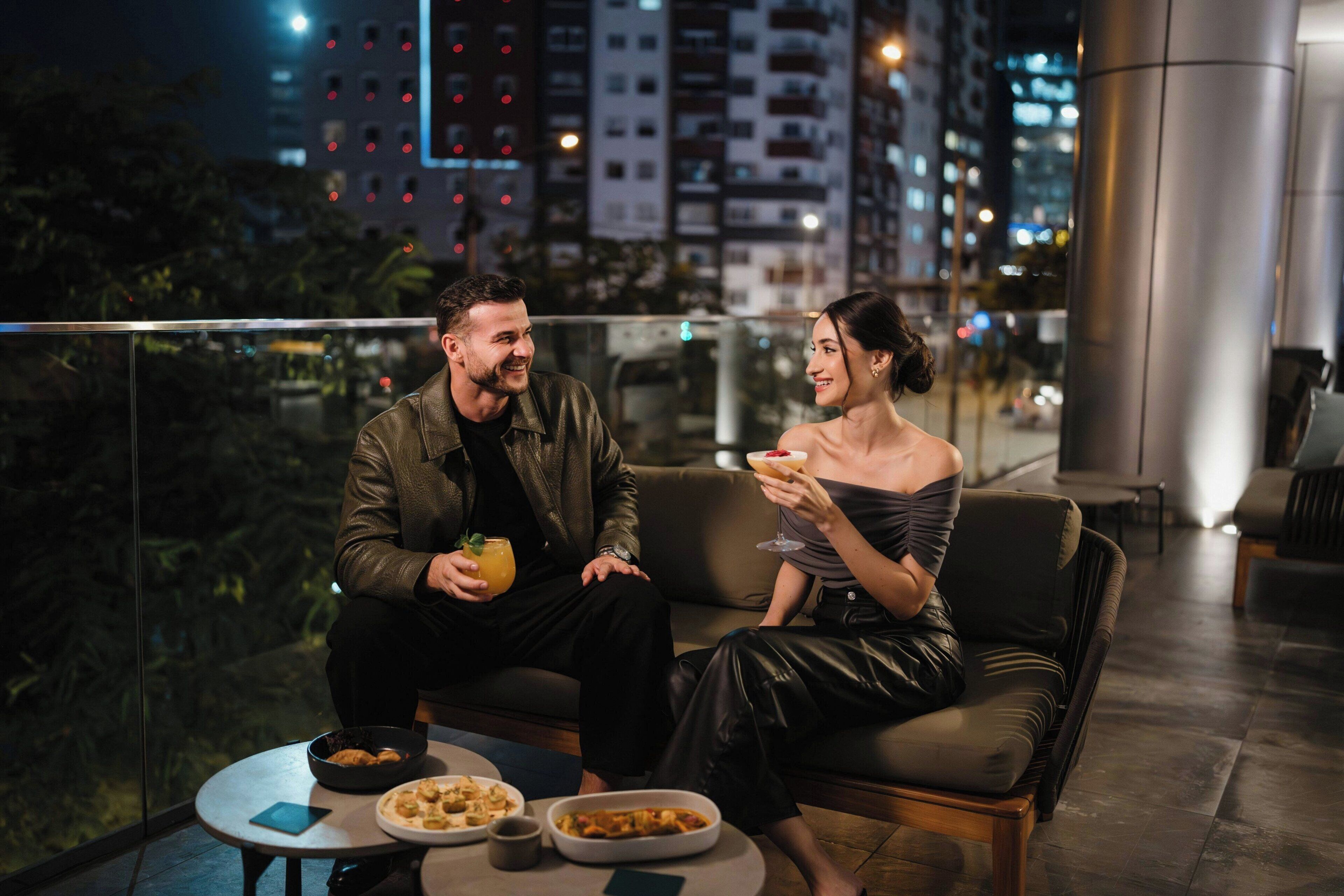 A couple seated on a balcony with a city view, a table with food and drinks, and a modern interior.