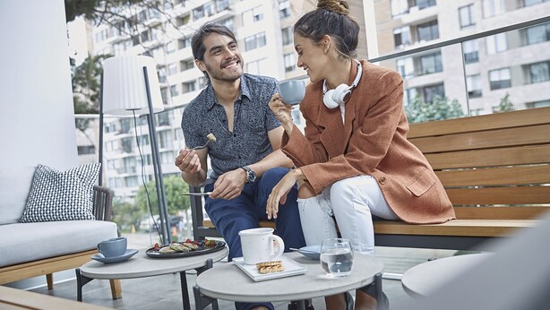 Una pareja sentada en un café al aire libre con vistas a la ciudad, mesa de comedor y una banca.