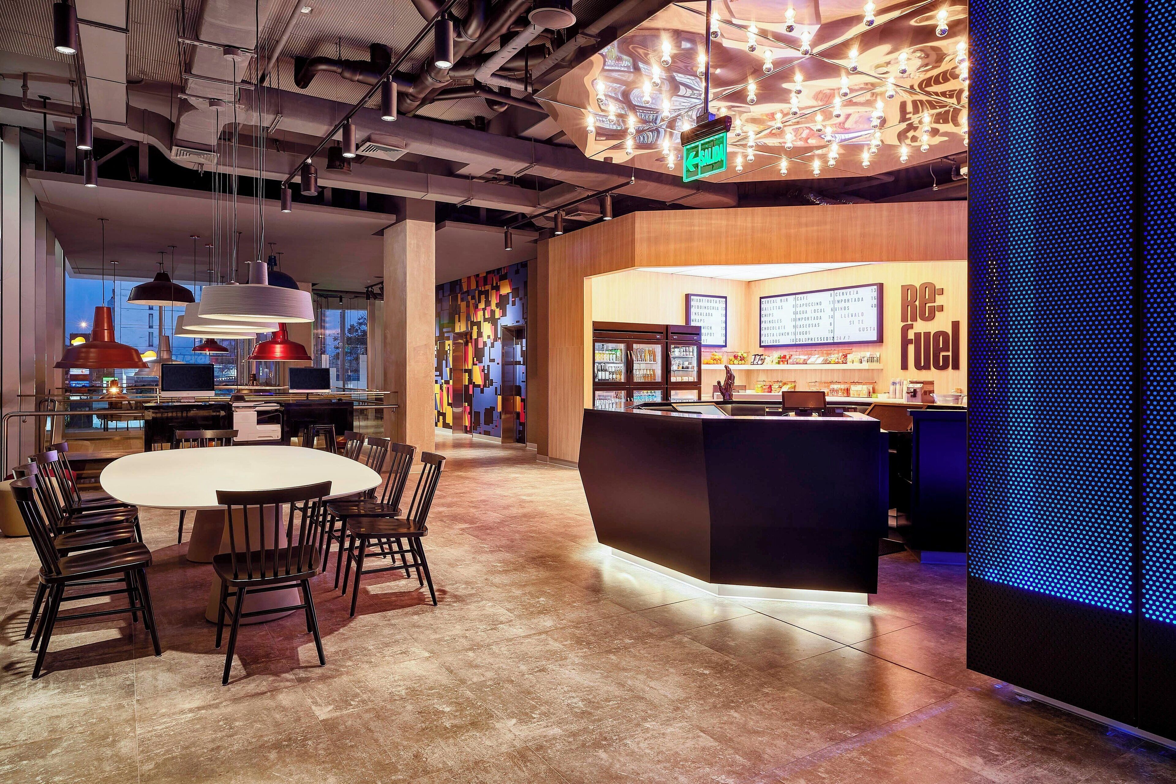 A modern restaurant with a central round table and chairs, a counter with a menu display, and a patterned ceiling.