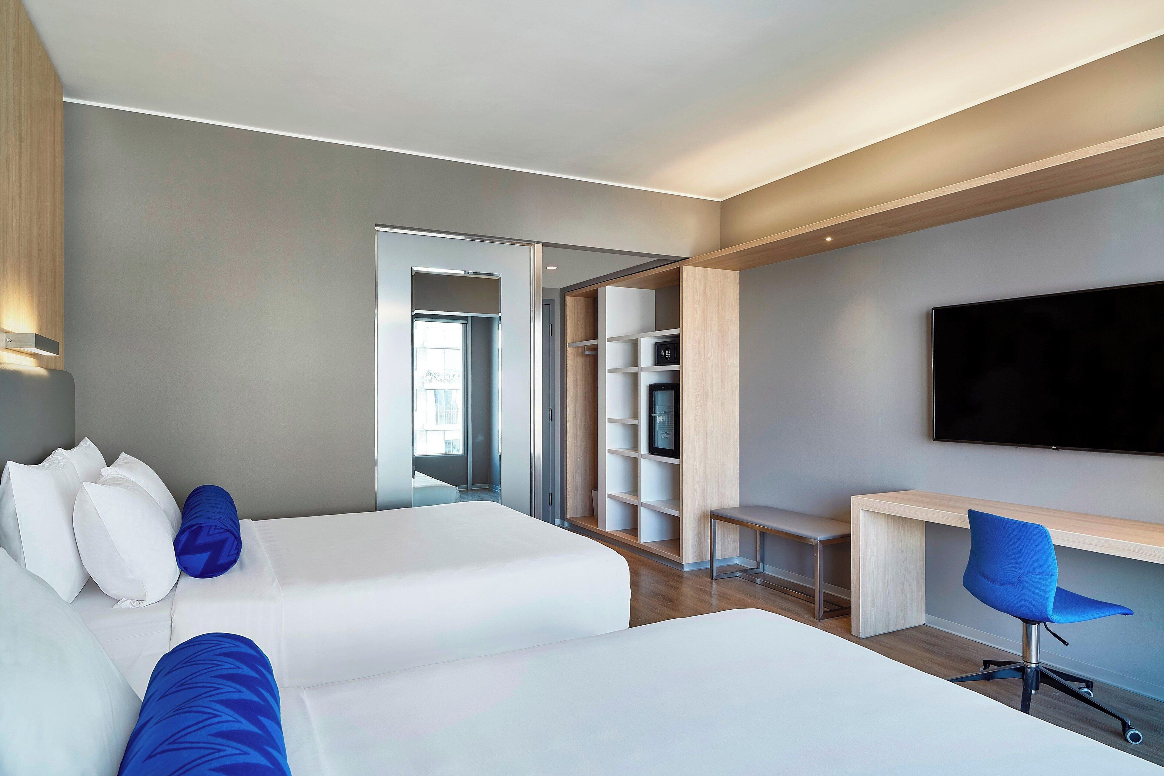 Habitación de hotel moderna con dos camas, un escritorio, una silla, un televisor y un armario.