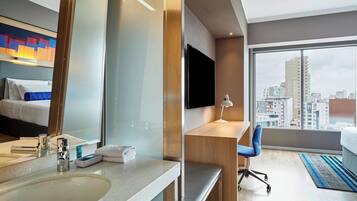 A hotel room with a bed, a desk, a computer, a TV, and a bathroom with a sink and mirror.