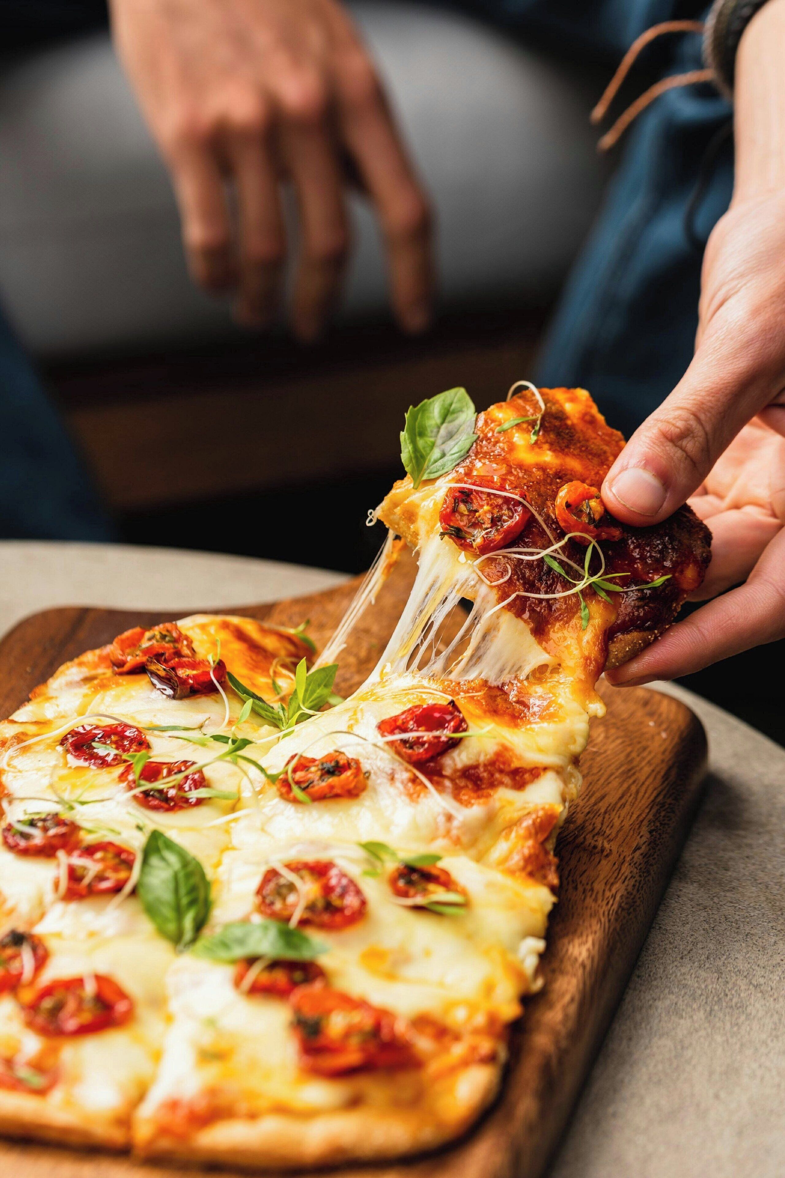 A hand pulling a slice of pizza with cheese and toppings from a wooden board.