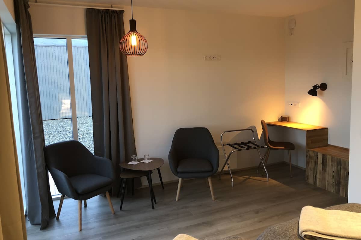 Una sala de estar moderna con un sofá, dos sillones, una mesita redonda y una mesa de centro de madera. Se aprecia un ventanal con cortinas, una lámpara colgante y una lámpara de pared.