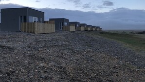 A row of modern, rectangular buildings with large windows, set on a gravel surface with a grassy area in the background. - Hrafnavellir Guest House (Höfn)
