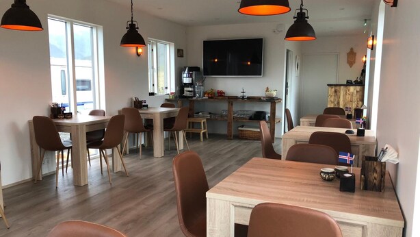 A modern dining area with wooden tables and brown chairs, a kitchen area with a large TV, and a view of the outdoors through large windows. - Hrafnavellir Guest House (Höfn)