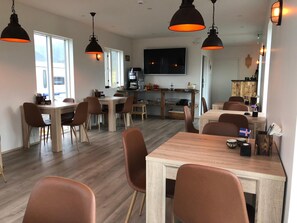 Een moderne eetruimte met houten tafels en bruine stoelen, een keuken met een grote televisie, en uitzicht op het buitenleven door grote ramen.