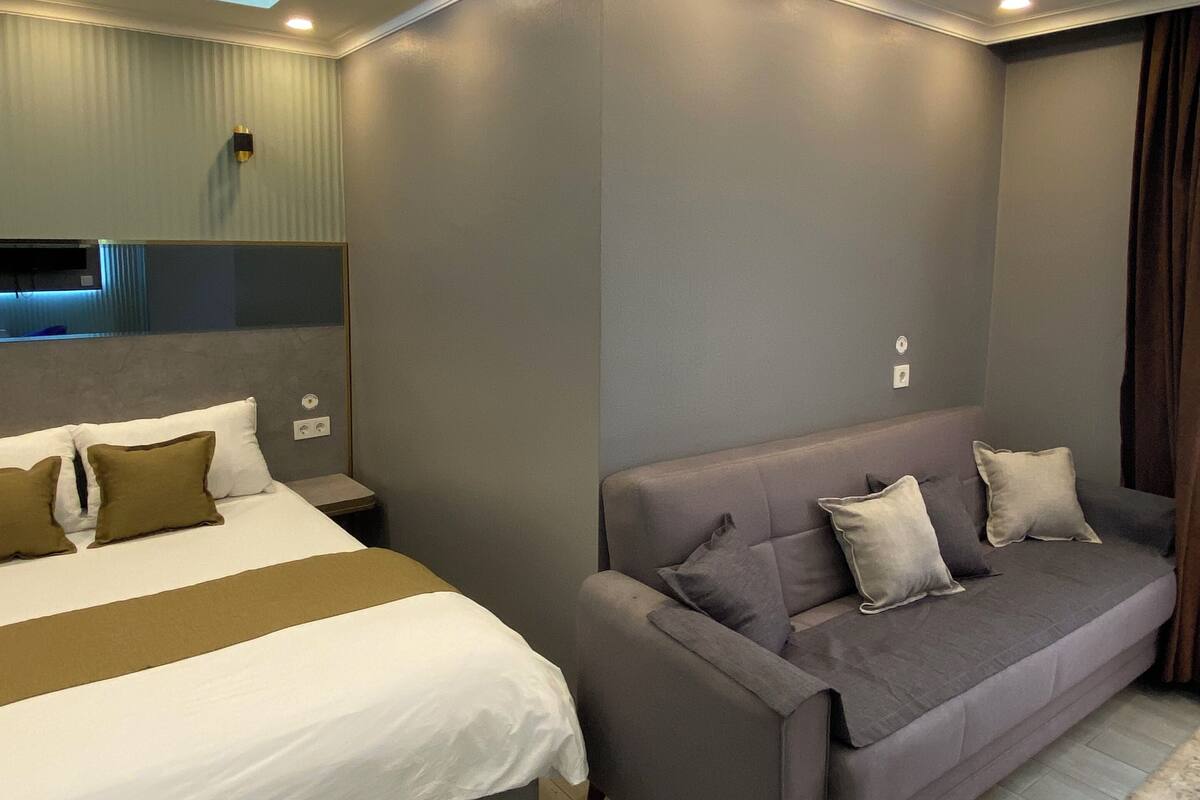 A modern hotel room with a bed, a sofa, and a ceiling with recessed lighting.