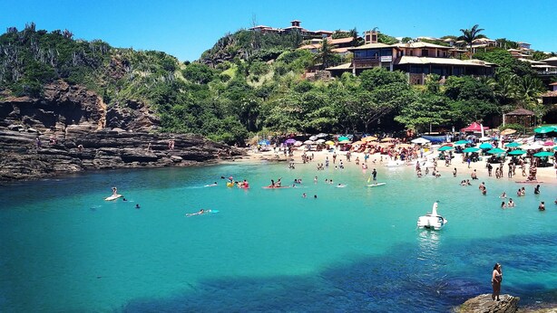 A beach with clear turquoise water, rocky shorelines, and a crowd of people enjoying the sun and sea. - Residence Solemare Buzios (Búzios)