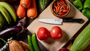 Diverse verse groenten en fruit, waaronder wortels, tomaten en paprika's, op een snijplank met een mes.