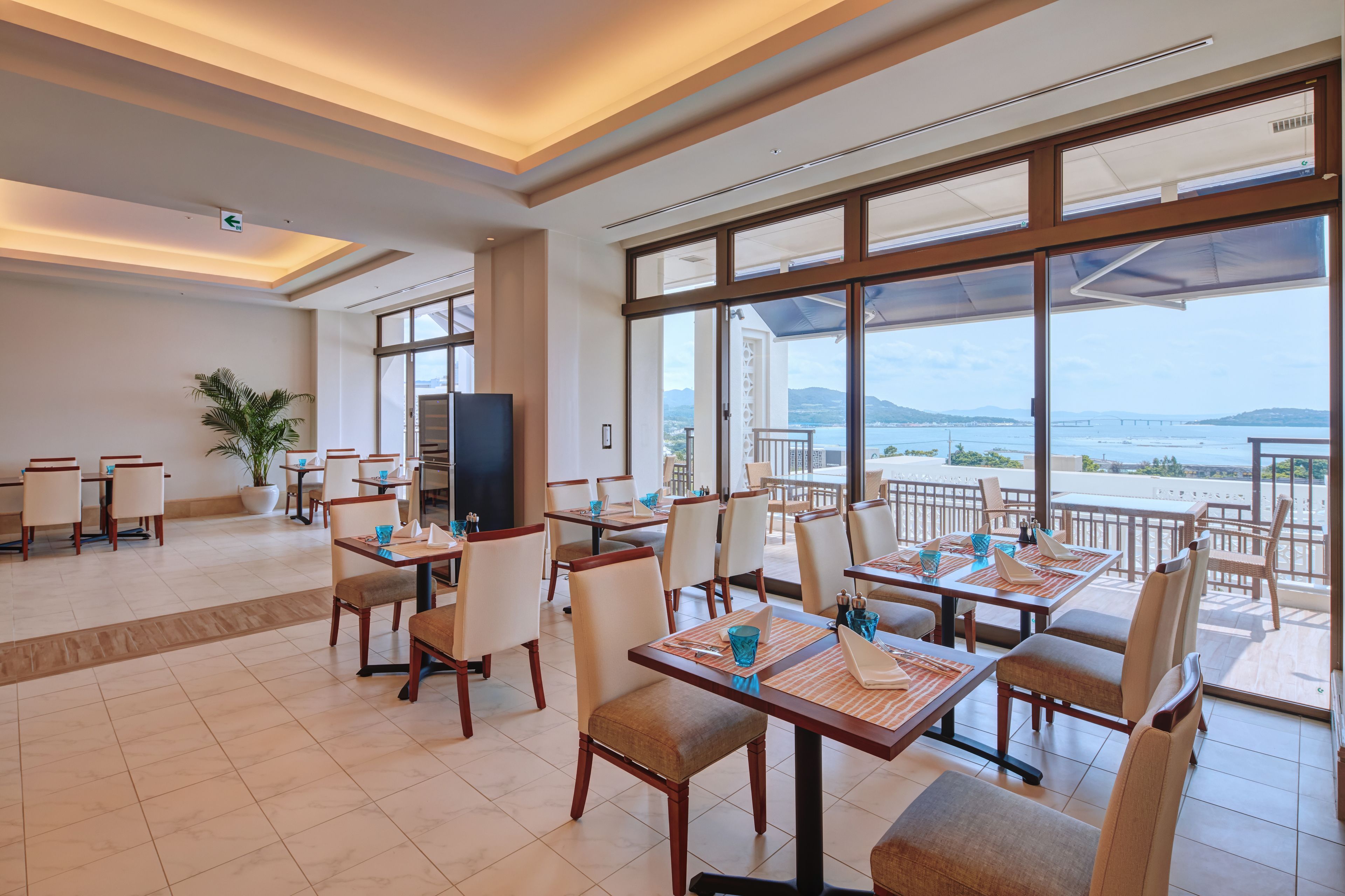 A spacious dining area with tables and chairs, large windows with a view of the sea, and a television on one wall.