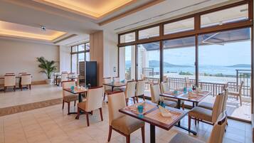 A spacious dining area with tables and chairs, large windows with a view of the sea, and a television on one wall.