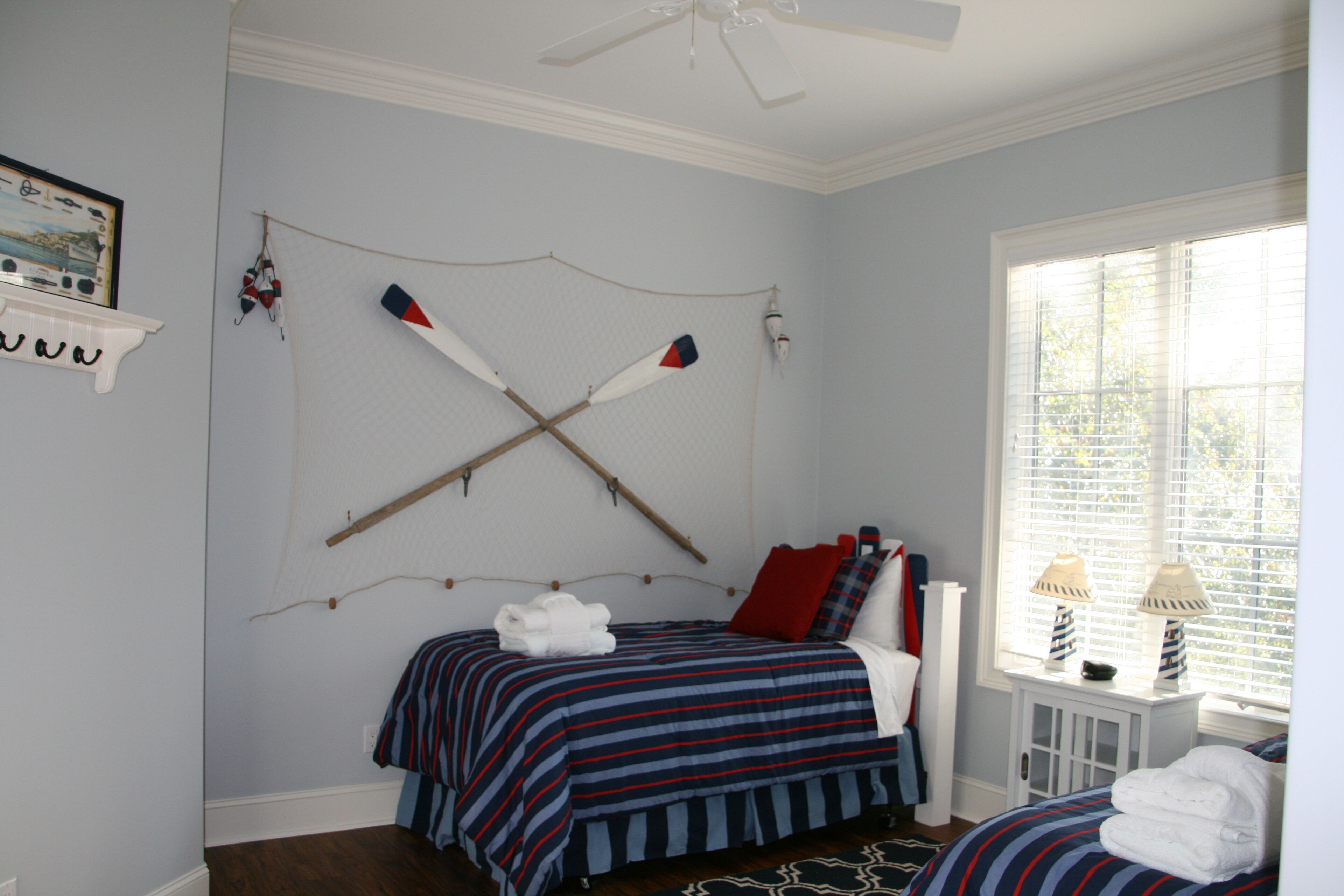 Un dormitorio con cama, ventana con persianas, ventilador de techo y repisa en la pared con un cuadro enmarcado.