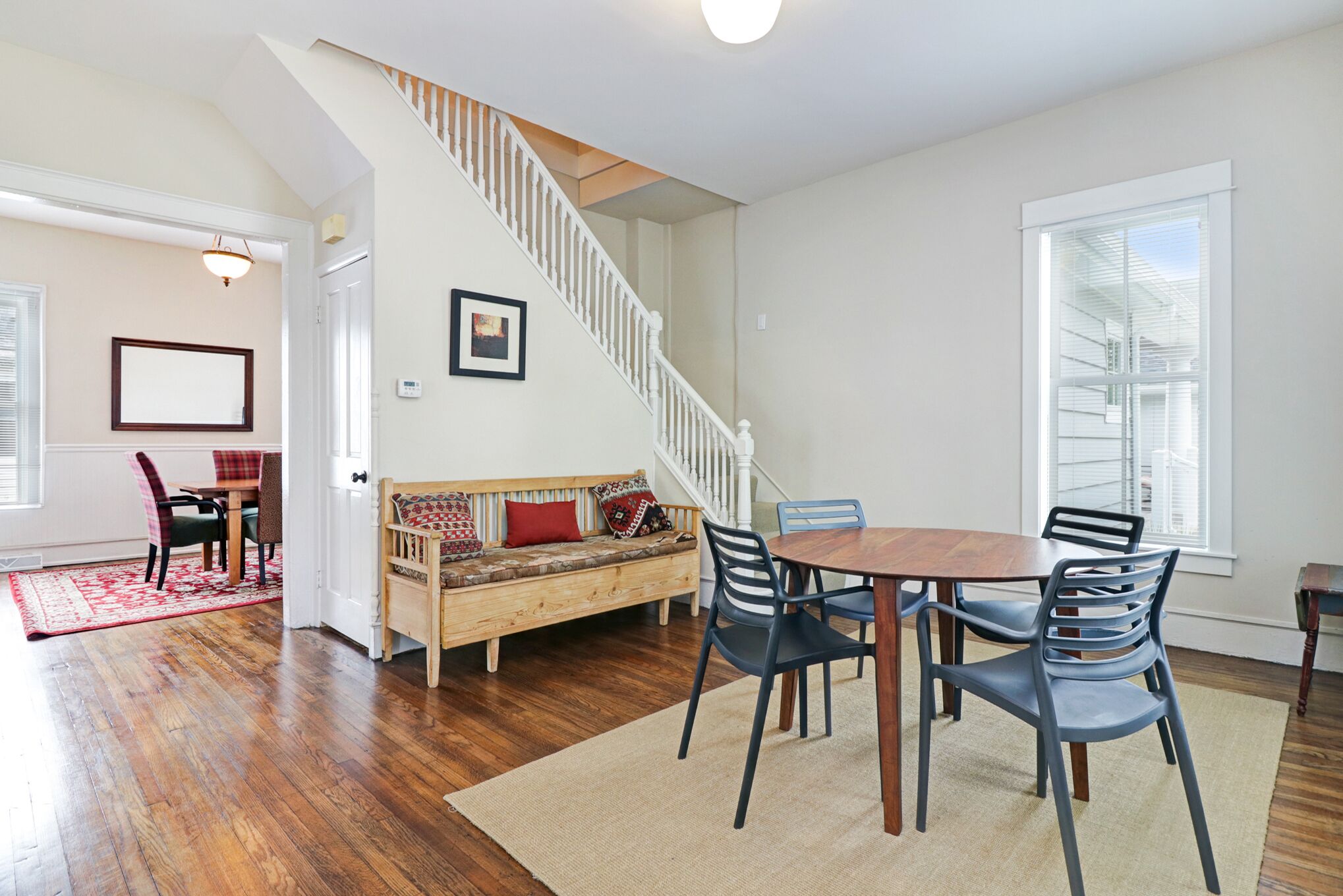 A spacious living area with hardwood flooring, a wooden dining table with chairs, a sofa with cushions, a staircase, and a large window providing natural light.