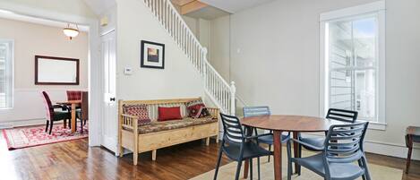 A spacious living area with hardwood flooring, a wooden dining table with chairs, a sofa with cushions, a staircase, and a large window providing natural light.