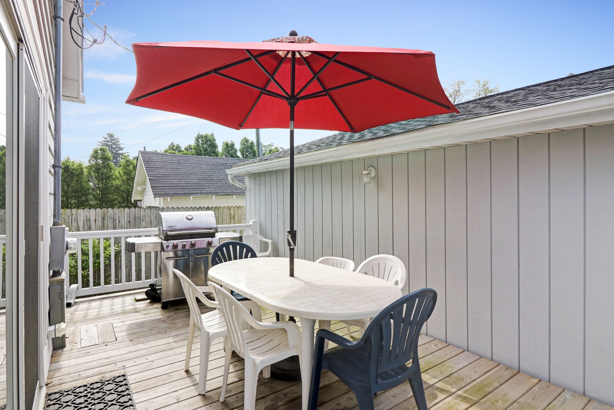 Una terraza con una mesa y sillas blancas, un paraguas rojo y una parrilla.