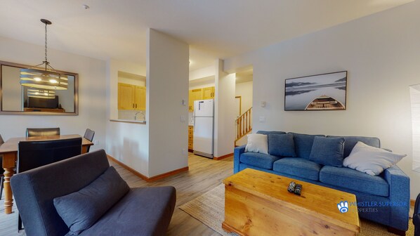 A modern living room with a blue sofa, a wooden coffee table, and a dining area with a table and chairs. - Stoney Creek Northstar 101 (Whistler)