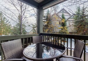A covered outdoor seating area with a round table and wicker chairs, overlooking a snowy landscape and a building with a distinctive roof. - Stoney Creek Northstar 101 (Whistler)