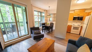 A spacious living area with a dining table, sofa, and wooden coffee table. A kitchen with wooden cabinets and appliances is visible in the background. - Central, Whistler Village Family Condo. Pool & Hot Tub, Free Parking & WiFi (Whistler)