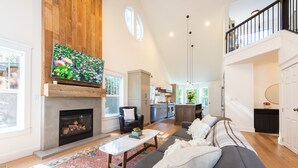 A spacious living room with a fireplace, a large TV mounted above it, a sofa, and a coffee table. - ~LARGE WESTSIDE HOME~ Walk to everything! Welcome to KINGSTON HOUSE (Bend)