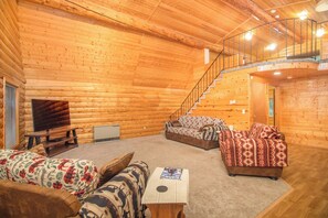 Interior de una cabaña de troncos con una escalera, dos sofás, un televisor y una mesa.