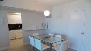 Un espace repas moderne avec une table en verre et des chaises blanches, une cuisine en arrière-plan et une porte blanche.