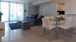 Un salon moderne avec une table à manger en verre, des chaises blanches, un canapé gris et un pouf à motifs.