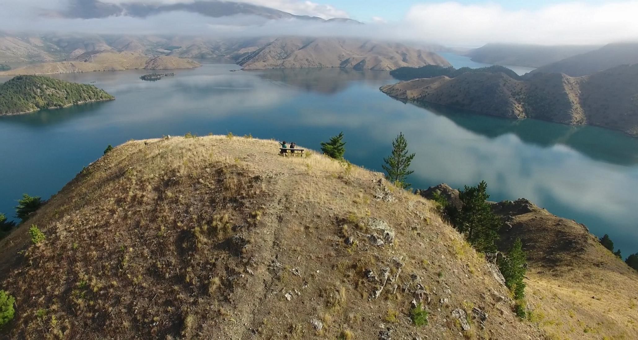 Paisaje montañoso con un lago y un pequeño grupo de personas sentadas en un banco.