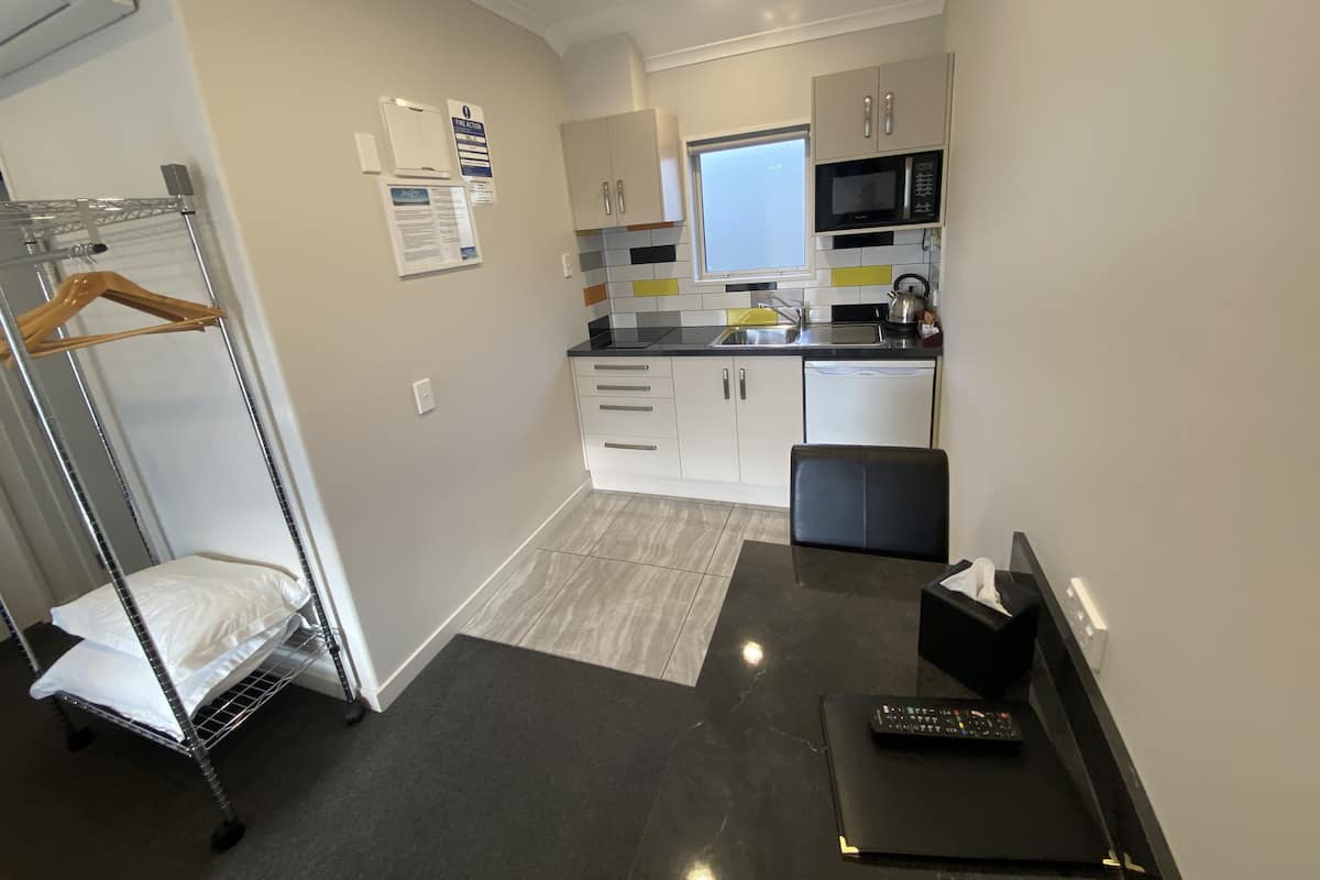 A compact kitchen with white cabinets, a microwave, and a sink. There's a small seating area with a black chair and a remote control on a table.