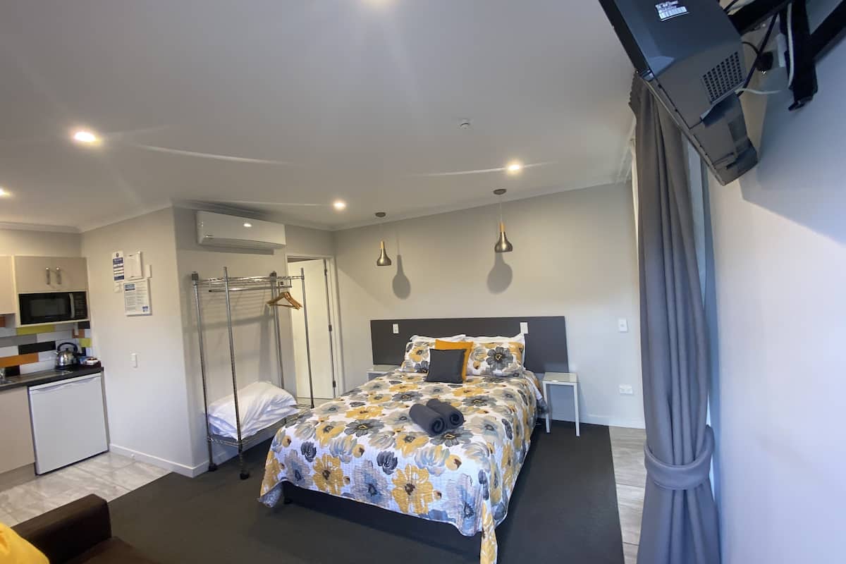 A bedroom with a bed, bedside table, and a desk. There is a kitchenette with a microwave and a refrigerator.