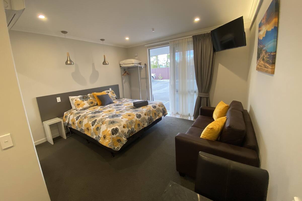A hotel room with a bed, a sofa, a TV, a window with curtains, and a door.