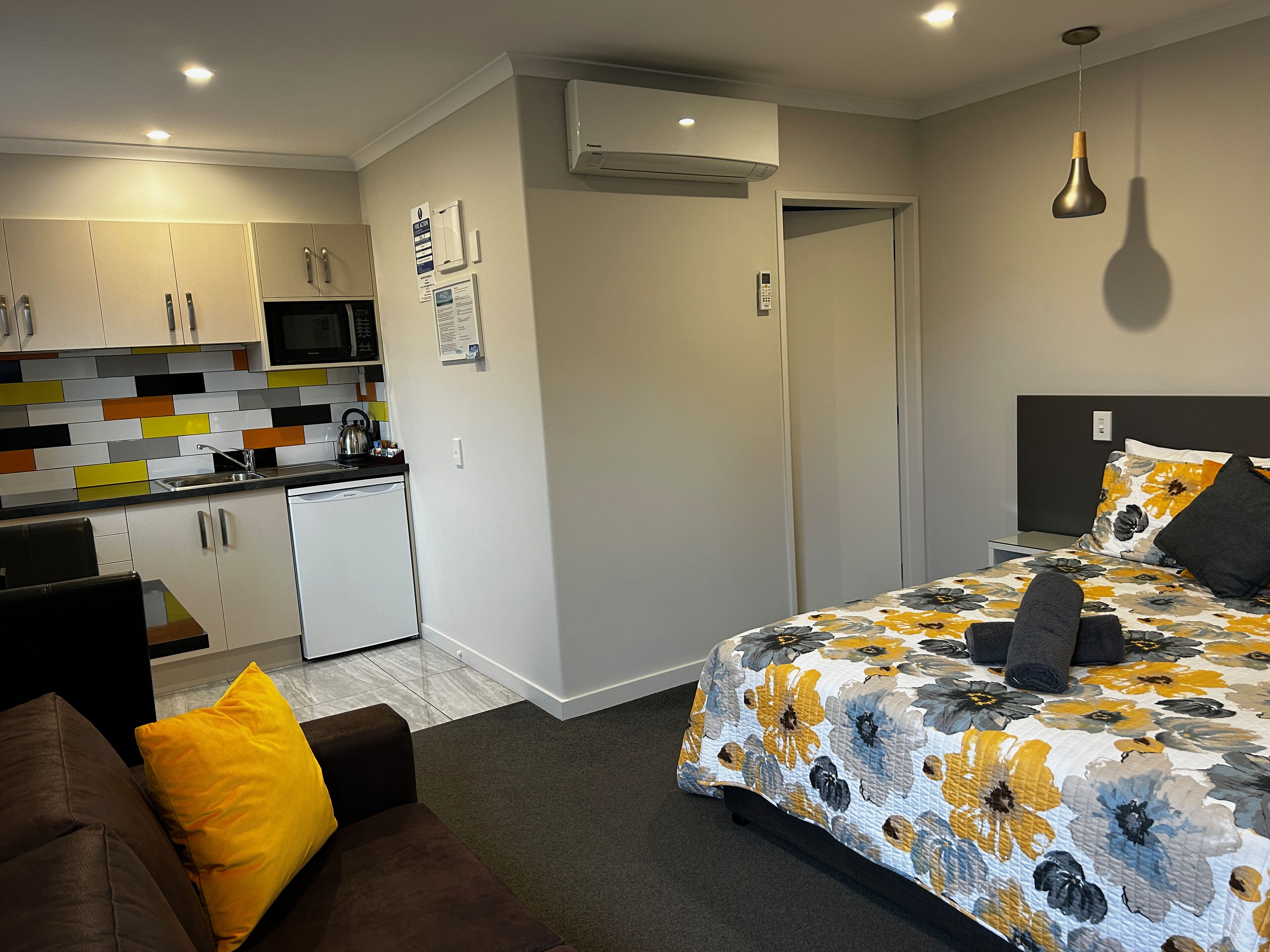 A compact hotel room with a kitchenette, a bed with floral bedding, a brown sofa with a yellow pillow, and a grey carpet.