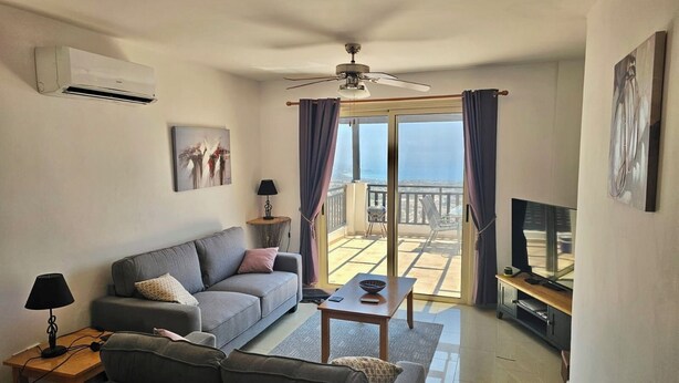 Ein modernes Wohnzimmer mit grauem Sofa, einem Couchtisch, einem Fernseher und einem Balkon mit Blick.
